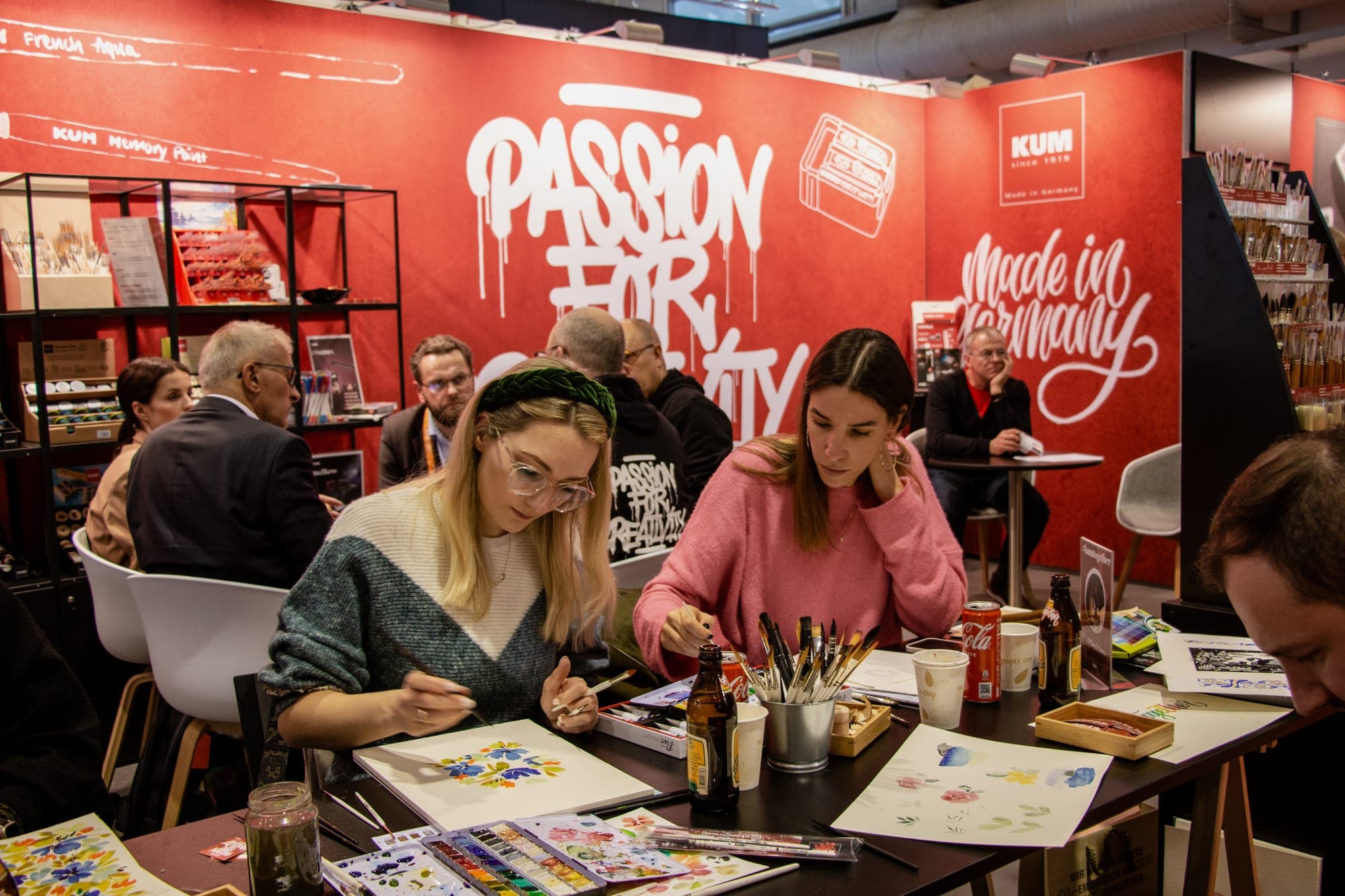 Zwei Frauen malen am Tisch vor der roten KUM-Wand mit Passion for Creativity