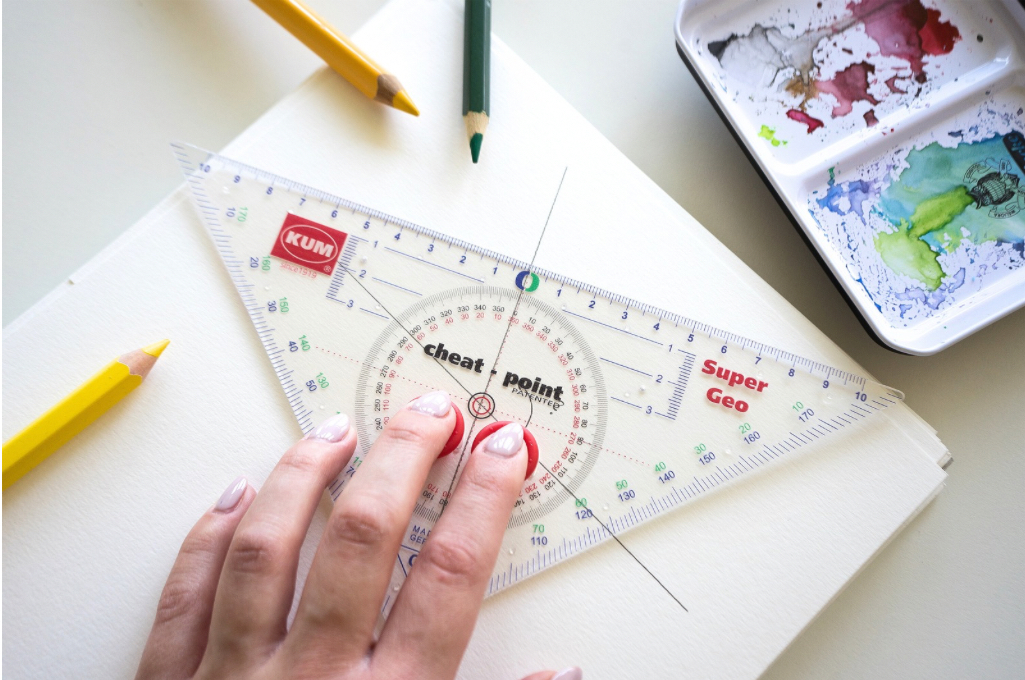 KUM Super Geo Geodreieck mit rotem Cheat-Point, von einer Hand auf einem Skizzenblock fixiert, daneben Buntstifte und ein Aquarellkasten