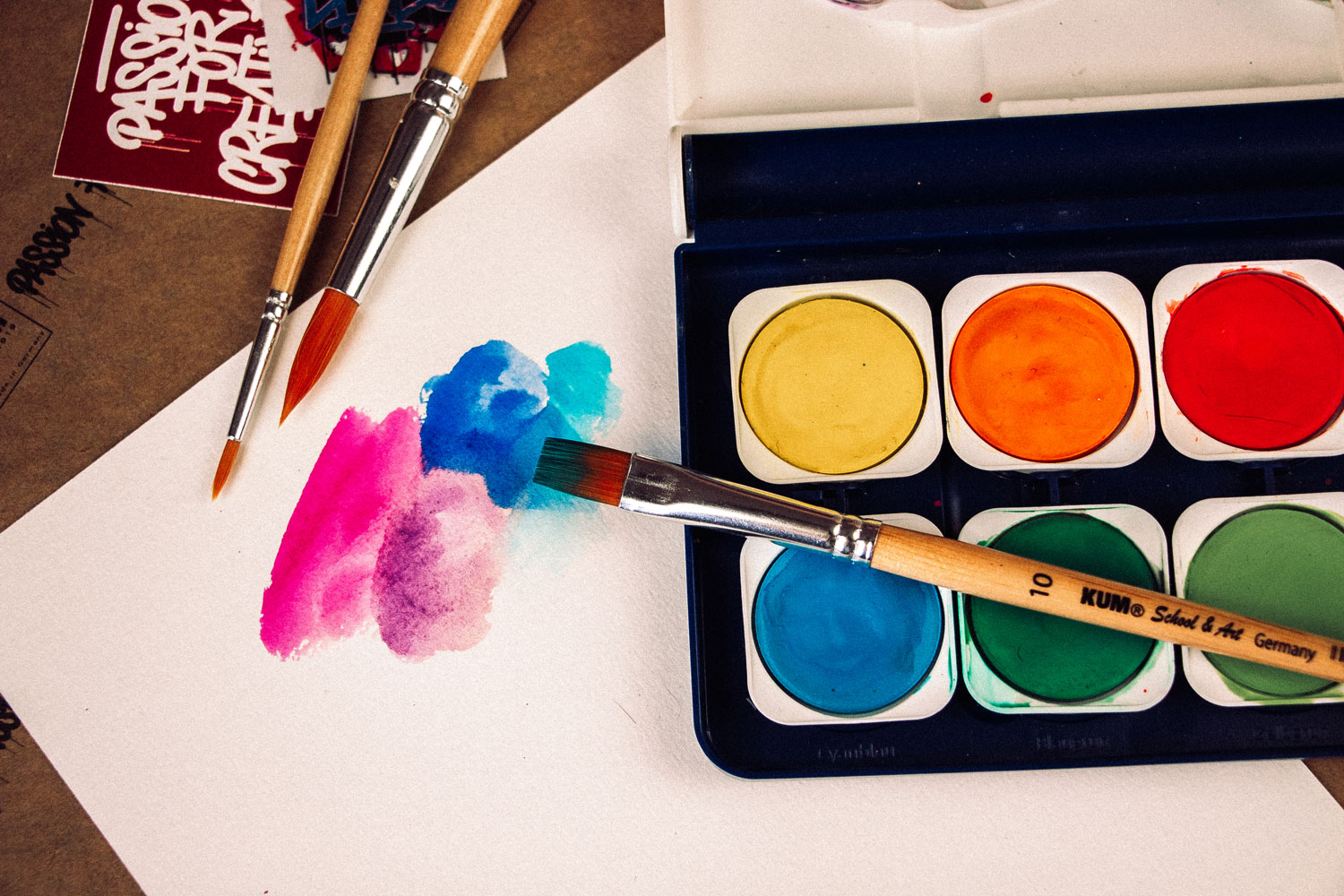 KUM Farbmalkasten mit 12 Farben, daneben ein School & Art Pinsel malt einen blau-pinken Aquarellverlauf