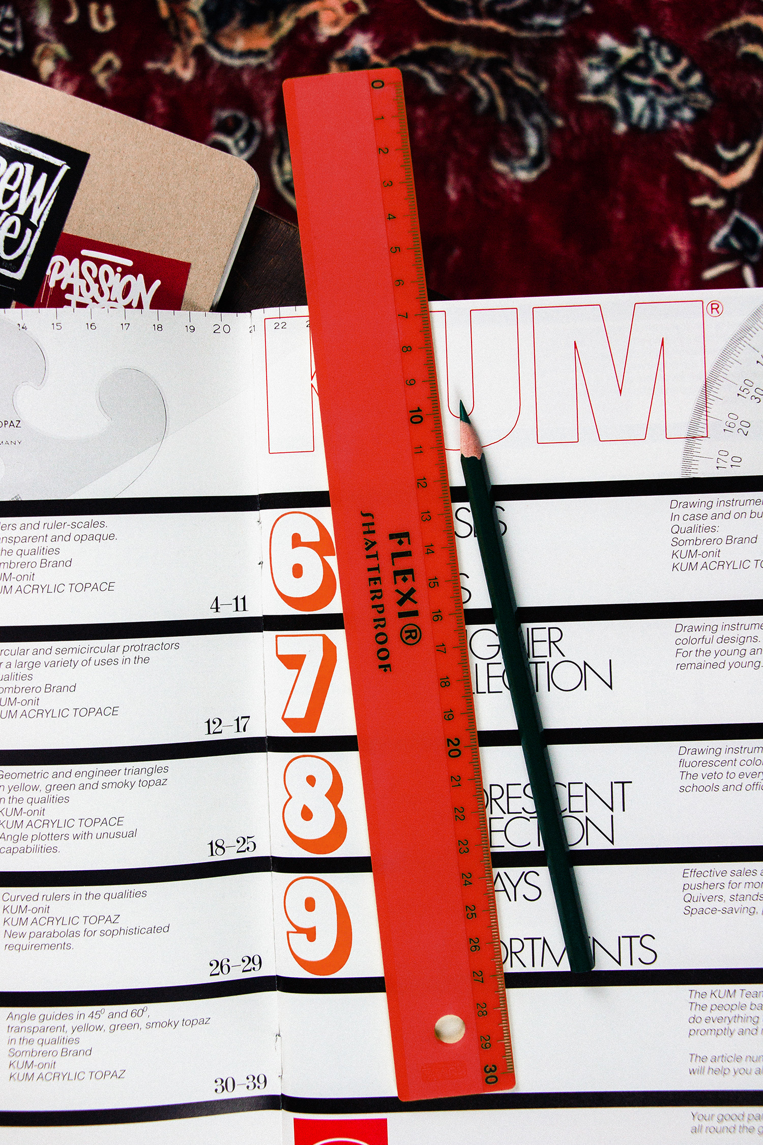 Rotes transparentes KUM Flexi Shatterproof Lineal auf einer aufgeschlagenen KUM-Katalogseite, daneben ein Bleistift
